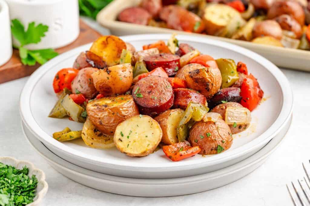 Plate of oven roasted sausage and vegetables with herbs; extra portion visible in baking dish behind.