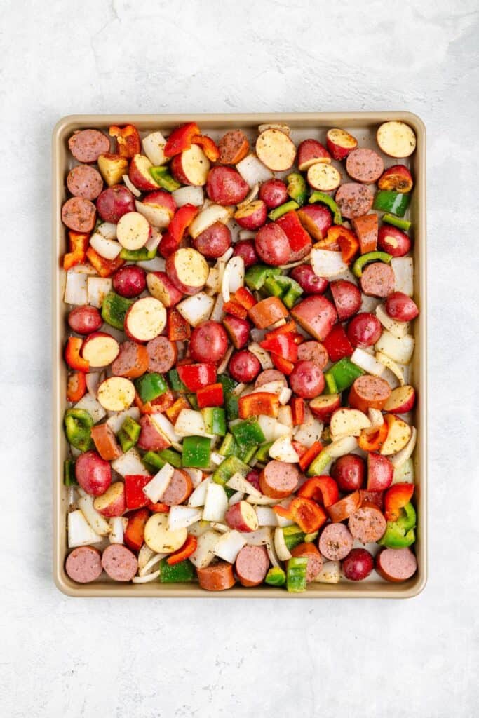 Chopped sausage, red potatoes, onions, and bell peppers on a baking sheet, seasoned for roasting.