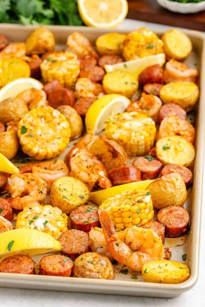 Sheet pan shrimp boil with baked, sausage slices, corn rounds, potato pieces, and lemon wedges arranged together.
