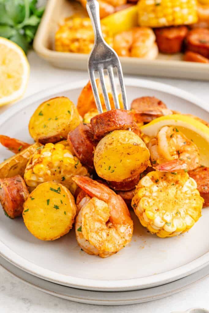 Plate of shrimp boil with sausage, corn, and potatoes; fork holds sausage. Lemon wedges and extra food in background.