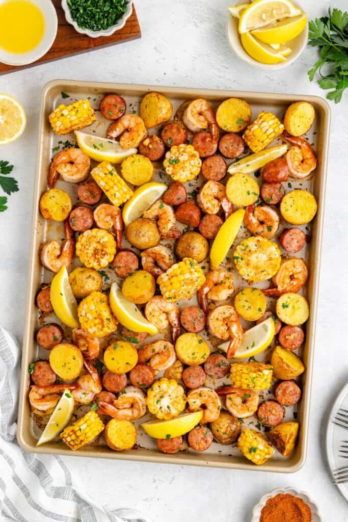 Shrimp boil sheet pan with roasted shrimp, sausage, corn, potatoes, and lemon wedges, garnished with chopped herbs.