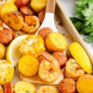Sheet pan shrimp boil with sausage, corn, potatoes, and lemon wedges; spatula lifting serving; parsley on side.