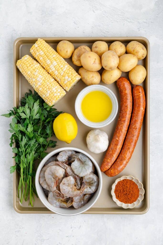Tray with raw shrimp, sausages, corn, baby potatoes, parsley, lemon, garlic, melted butter, seasoning, and paprika.