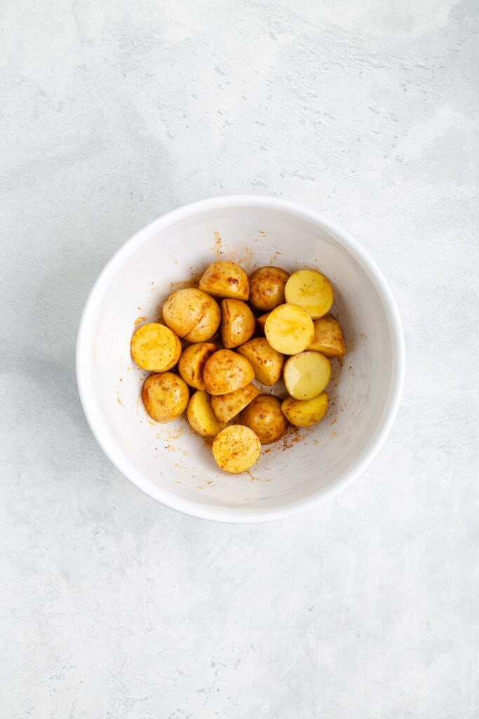 White bowl of halved baby potatoes coated in spices on a light gray surface.