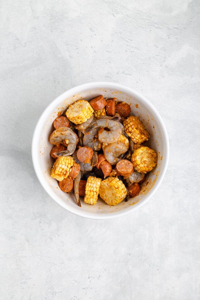 A white bowl with raw shrimp, sliced sausage, and corn on the cob, all seasoned and uncooked.