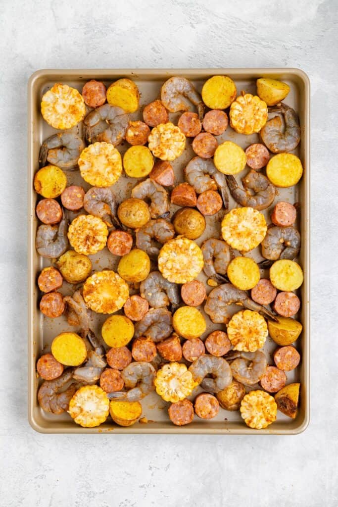 Raw shrimp, sausage slices, halved potatoes, and corn rounds arranged evenly on a baking sheet for roasting.