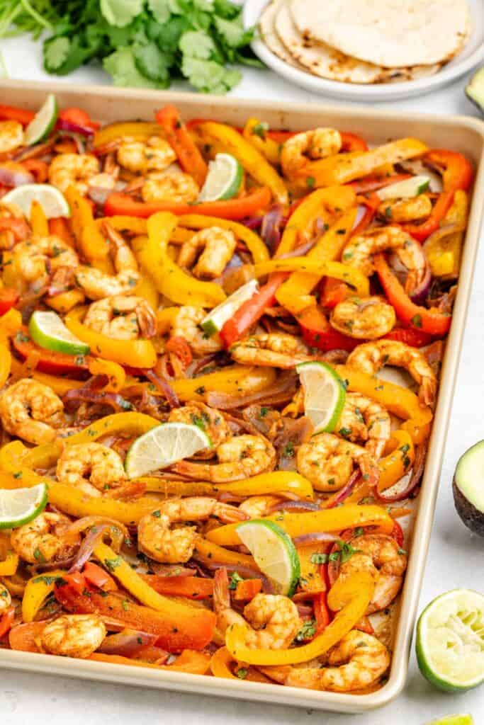 Sheet pan shrimp fajitas, bell peppers, onions, and lime; tortillas and fresh cilantro in background.