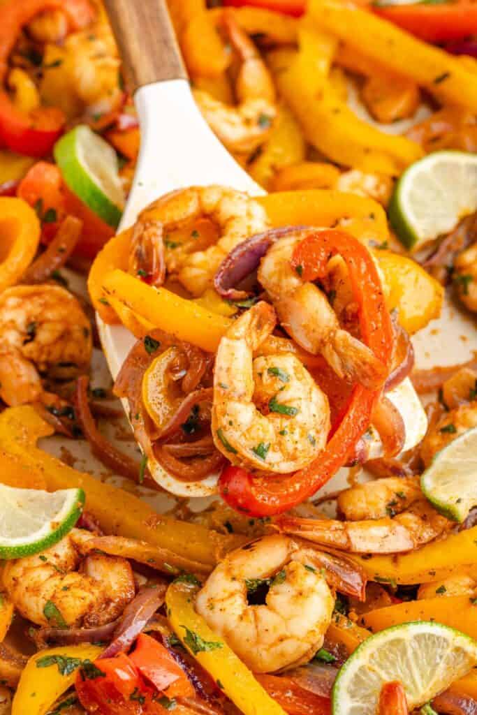Serving spoon with cooked shrimp fajitas, yellow and red bell peppers, red onions, and lime wedges.