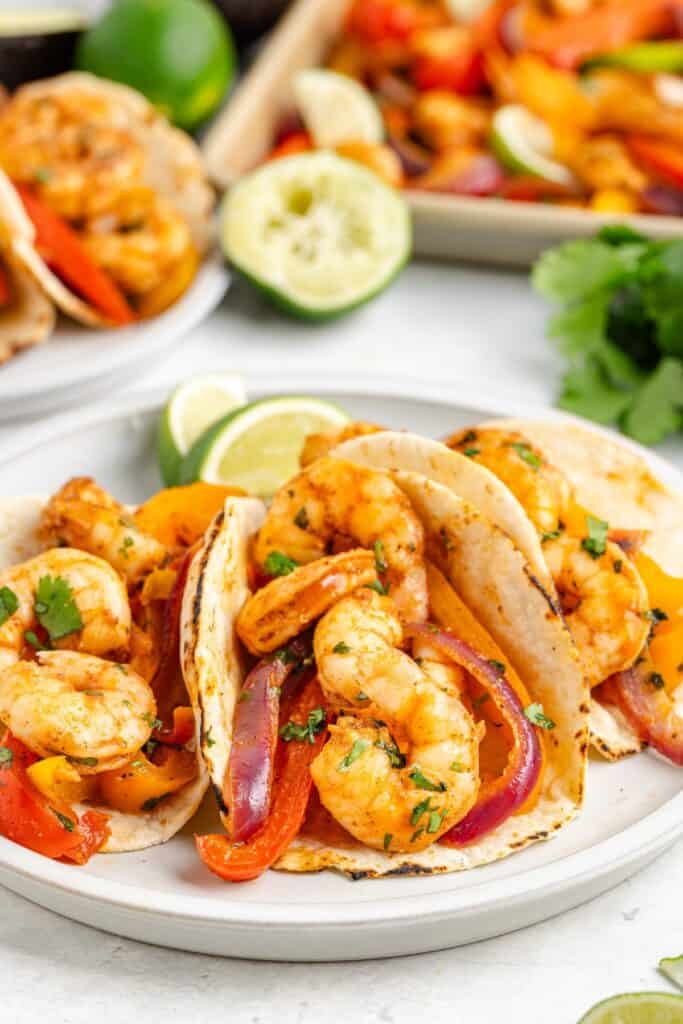 Three shrimp tacos with grilled peppers and onions, topped with cilantro, on a plate with lime wedges.