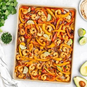 Sheet pan shrimp fajitas with colorful bell peppers with limes, surrounded by cilantro, avocado, tortillas.