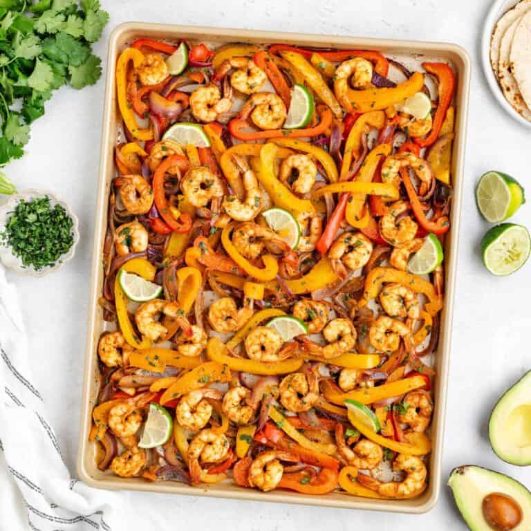 Sheet pan shrimp fajitas with colorful bell peppers with limes, surrounded by cilantro, avocado, tortillas.
