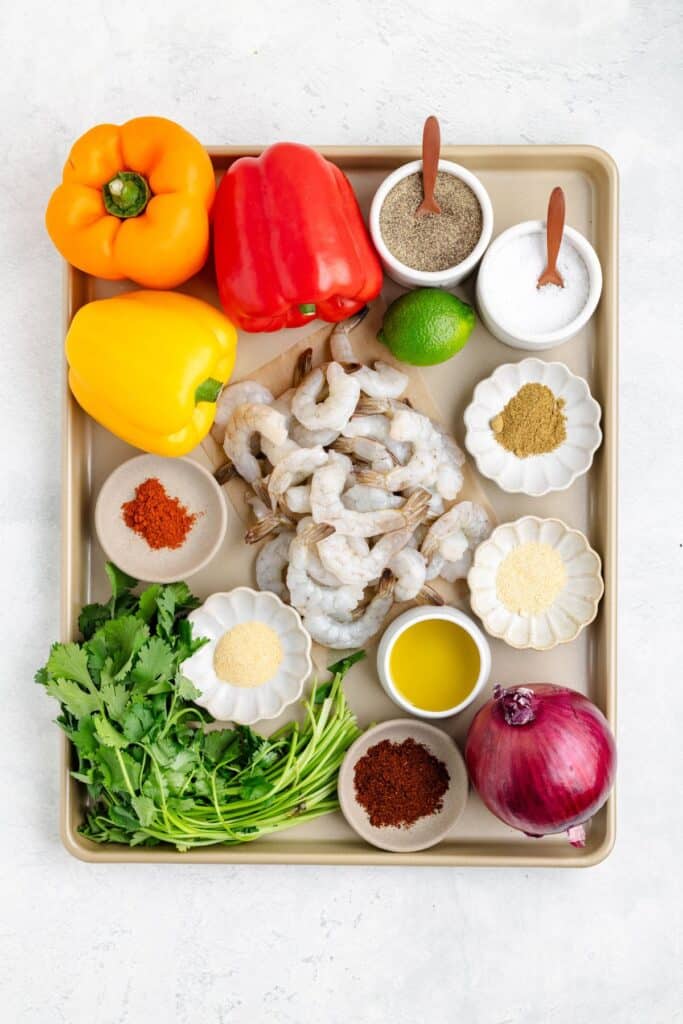 Tray with raw shrimp, colorful bell peppers, red onion, cilantro, lime, olive oil, and assorted spices.