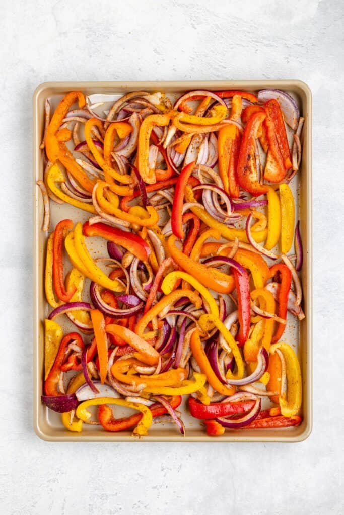 Sliced red, yellow, orange bell peppers and red onions spread on a baking sheet, ready for roasting.