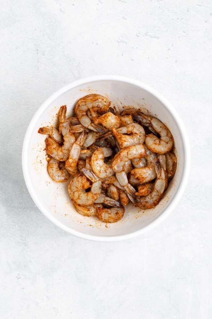 White bowl of raw shrimp coated in seasoning on a light-colored surface.