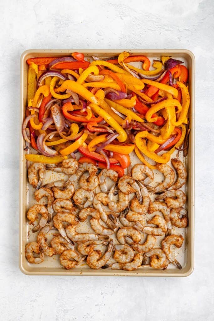 A baking sheet with seasoned shrimp beside sliced bell peppers and red onion, ready for cooking.