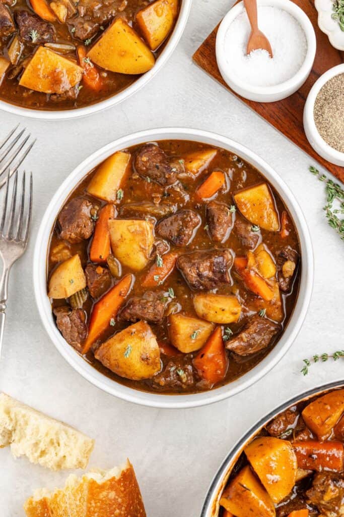 Bowl of easy beef stew with potatoes, carrots, bread, utensils, and small dishes of seasoning on table.