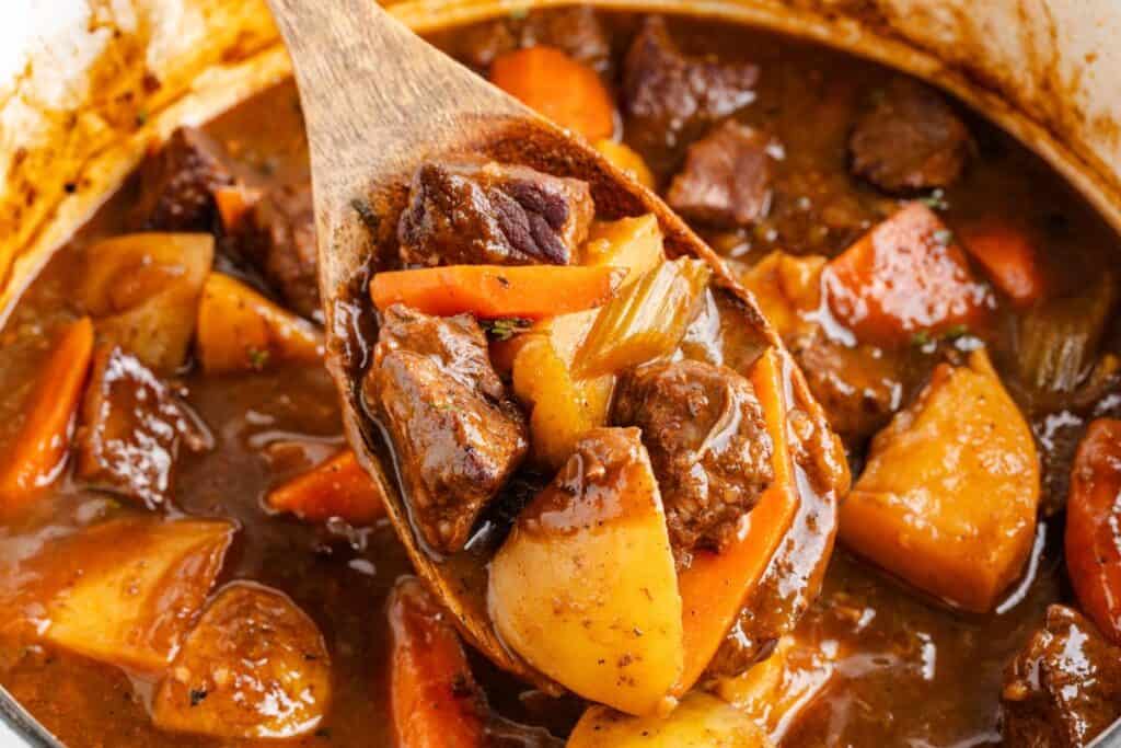 Wooden spoon with beef stew, potatoes, carrots, and brown broth held above a pot.