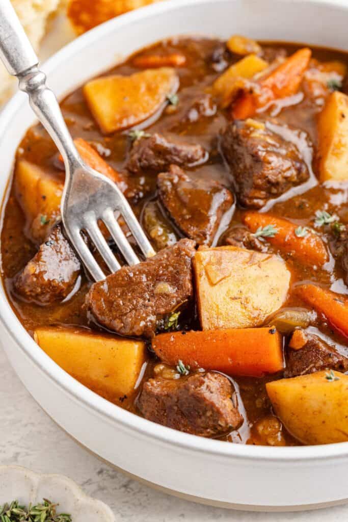 Bowl of stovetop beef stew with beef, potatoes, carrots, green beans in brown broth; fork resting on top.