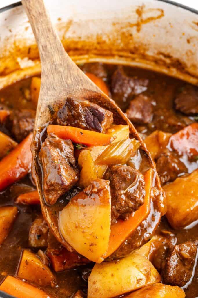 Wooden spoon holding beef stew with beef, potatoes, carrots, onions, and thick brown broth.