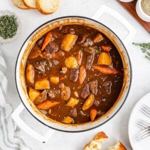 Pot of stovetop beef stew with meat, potatoes, carrots; bread, herbs, and utensils on a white surface.