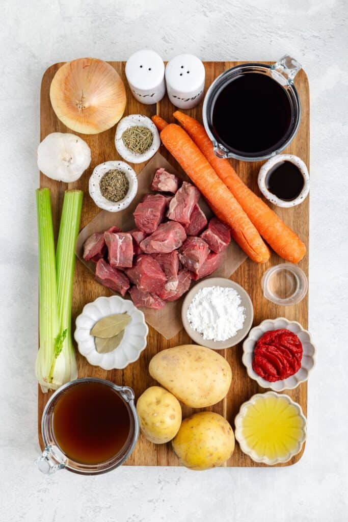 Wooden board with cubed beef, chopped stew vegetables, and small bowls of spices, wine, broth, and oil.