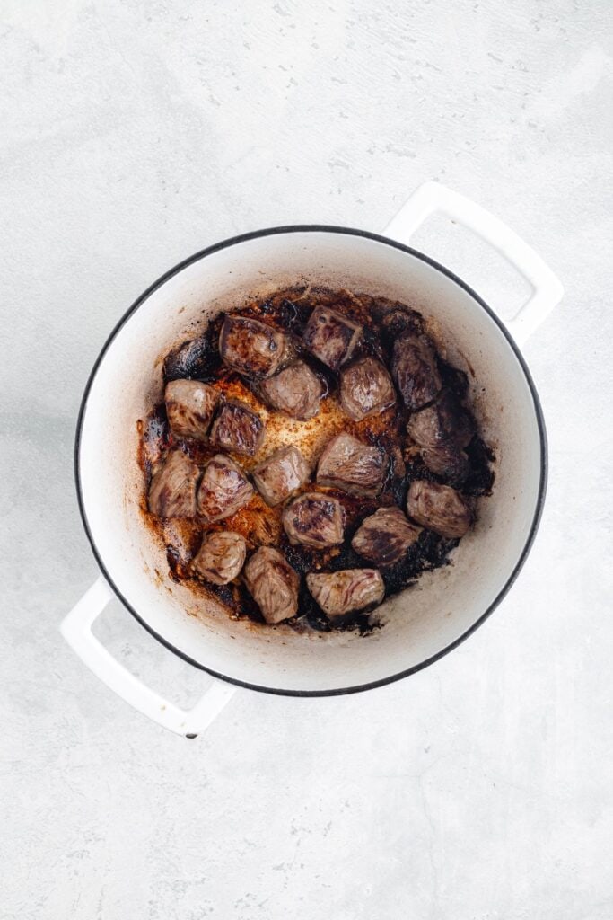 Browned beef cubes with caramelized, seared edges in a white Dutch oven on a light-colored surface.