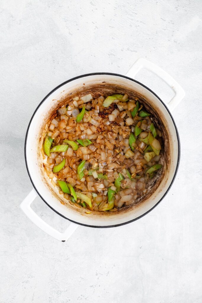 White pot with sautéed diced onions and celery, browned bits visible on sides, on light-colored surface.
