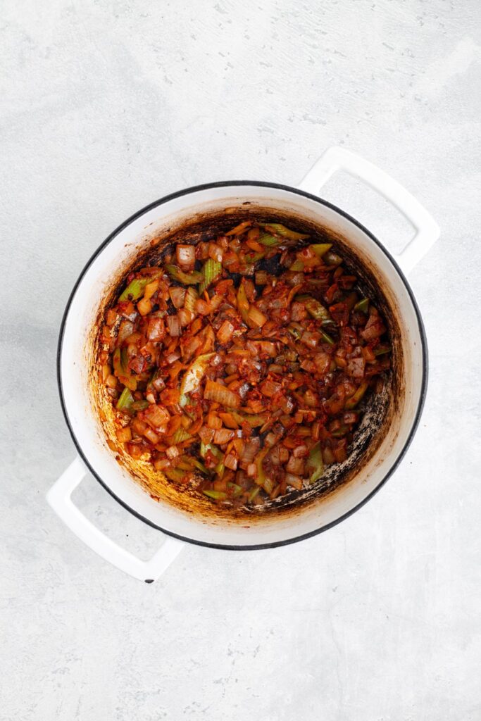 White pot with sautéed onions, celery, and tomatoes, partially caramelized, on a light gray surface.