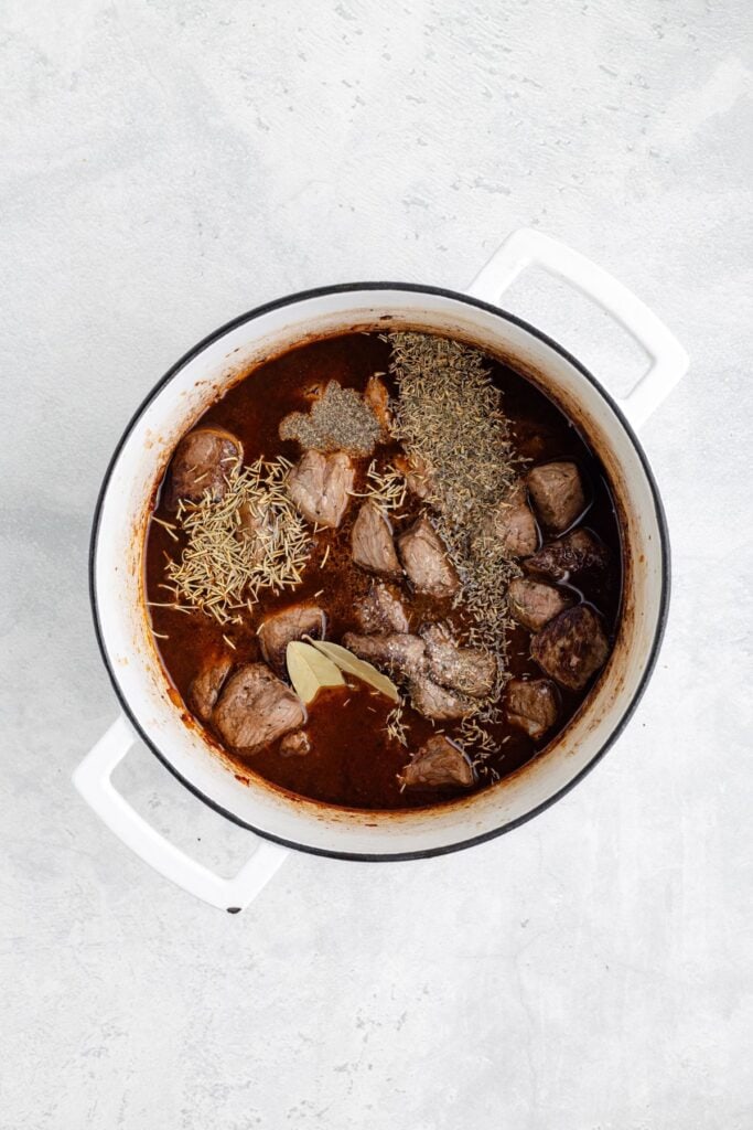 White pot with beef chunks, brown stew, and assorted dried herbs including bay leaves, viewed from above.