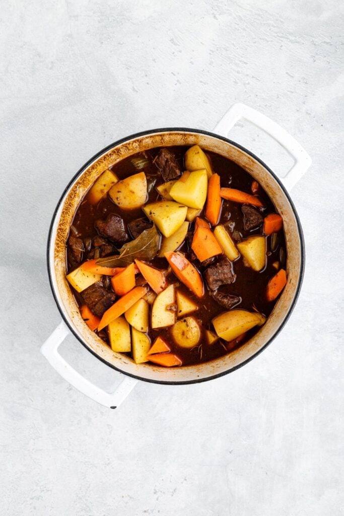 White pot filled with beef stew containing beef chunks, potatoes, and carrots in brown broth on light background.