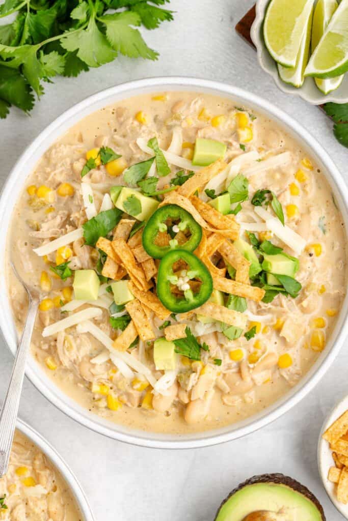 A bowl of creamy white chicken chili with jalapeños, tortilla strips, cheese, avocado, cilantro, and lime wedges.