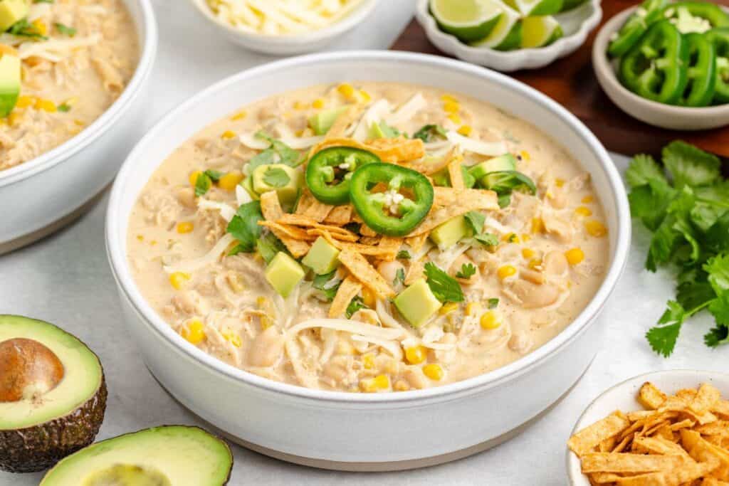 Bowl of stovetop white chicken chili topped with jalapeños, avocado, cheese, tortilla strips, cilantro; corn and avocado beside.