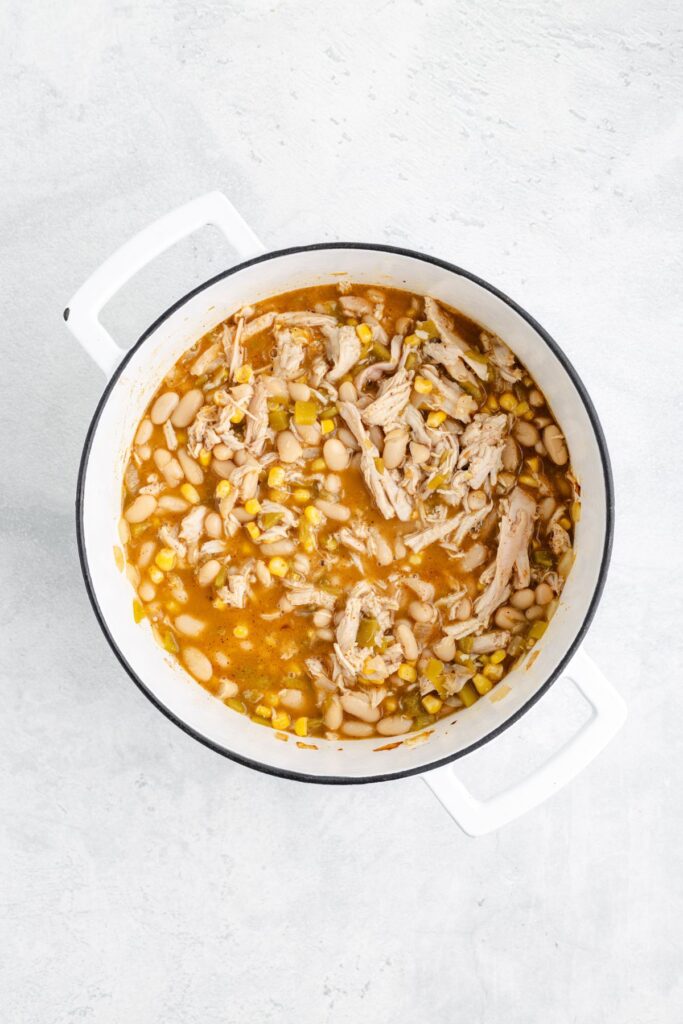 White pot with white bean chicken chili, featuring shredded chicken, white beans, corn, and broth on light surface.