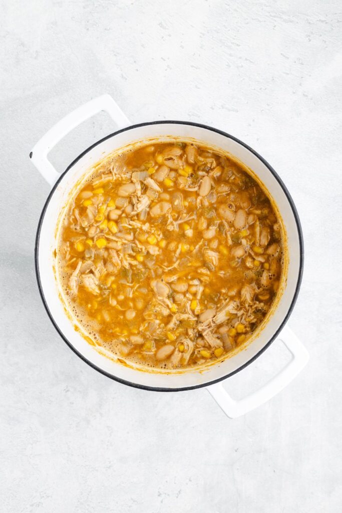White pot with shredded chicken, beans, and corn in golden broth seen from above on light background.