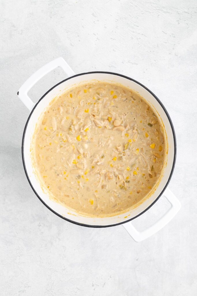White pot of creamy chicken chili with corn, beans, and shredded chicken on a light gray surface.