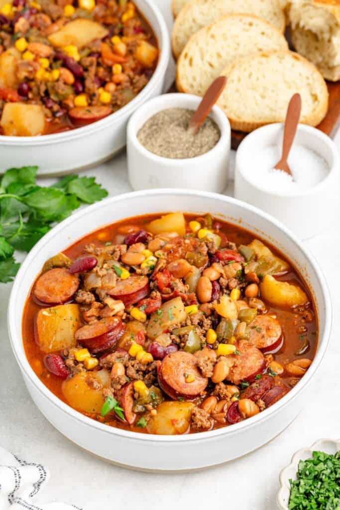 Bowl of hearty cowboy stew with sausage, beans, veggies, and sliced bread, salt, pepper, herbs on the side.