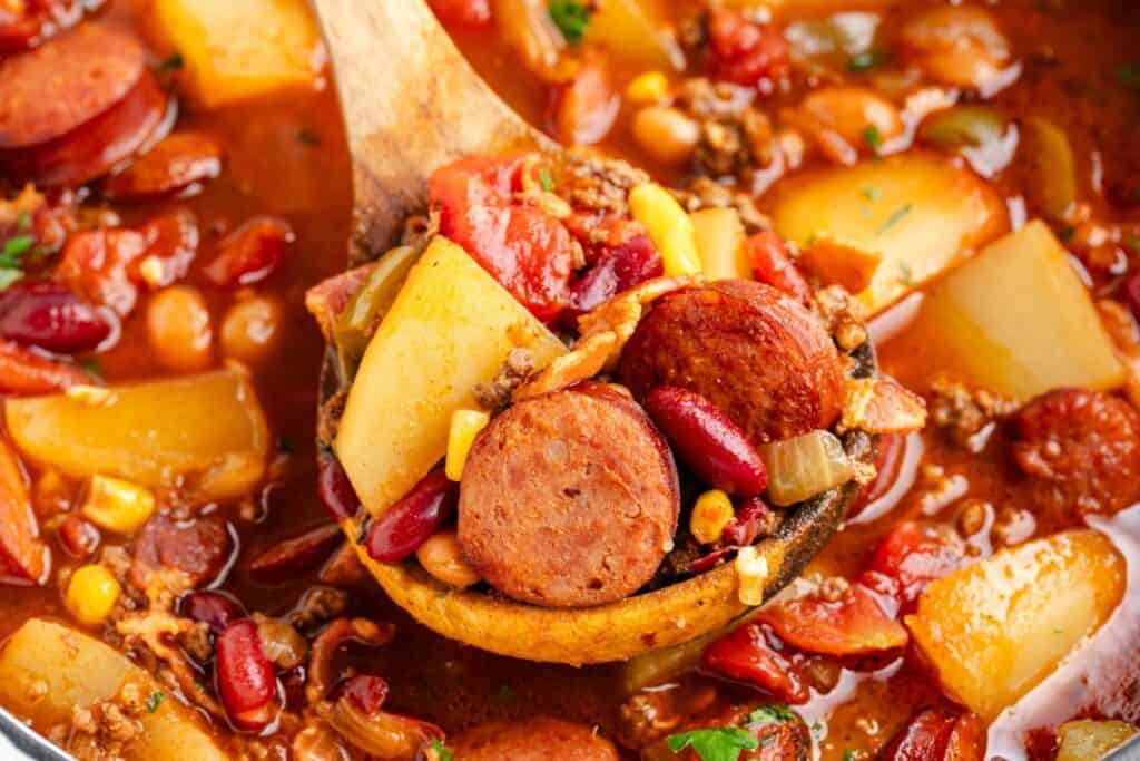 A wooden ladle lifts Texas cowboy stew with sausage, potatoes, beans, corn, ground meat, and tomatoes in rich broth.