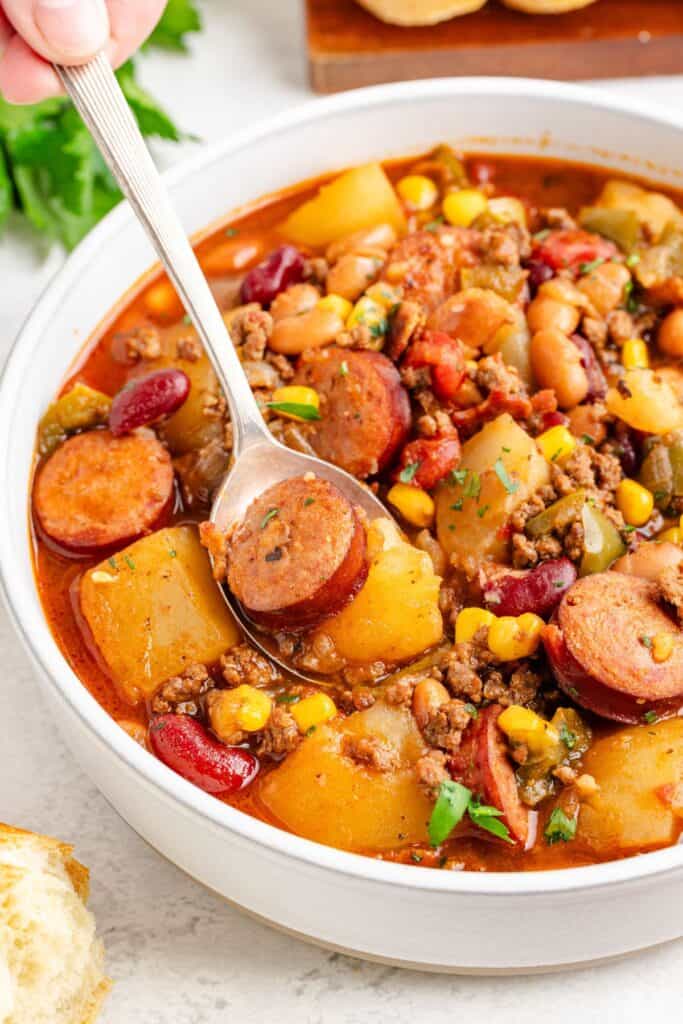 A bowl of cowboy stew with sausage, ground meat, beans, potatoes, corn, vegetables; spoon lifting a serving.