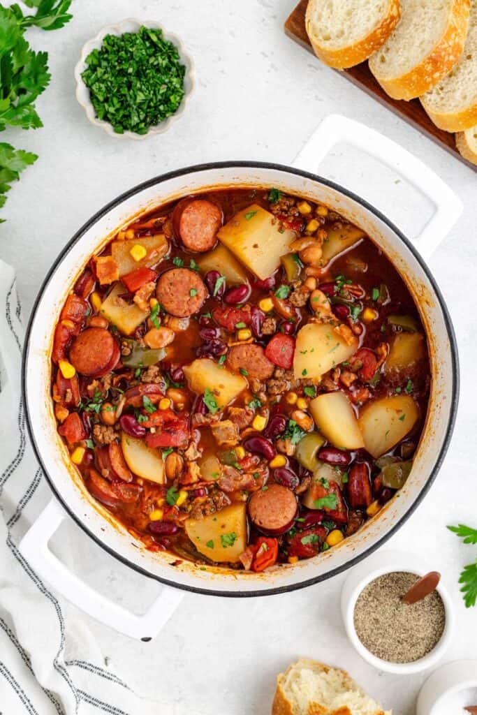 White pot of sausage and vegetable stew with potatoes, beans, corn; sliced bread and herbs surround on white surface.