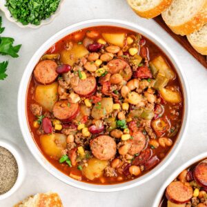 Hearty cowboy stew with sausage, beans, potatoes, corn, and tomatoes in a bowl, bread slices and parsley nearby.