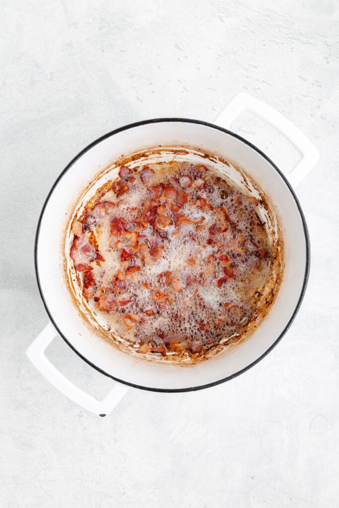 White pot on light surface with browned, chopped bacon pieces and rendered fat; some browned residue visible.