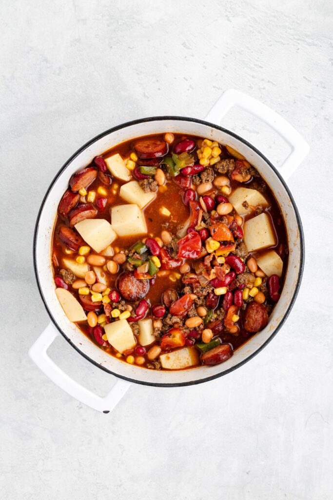 White pot of mixed vegetable, bean, and sausage stew in red broth, viewed from above on a light surface.