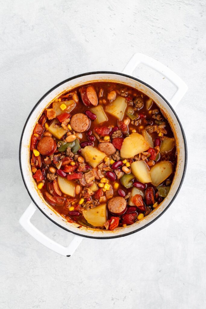 White pot with chunky stew of sausage, potatoes, beans, corn, tomatoes, and vegetables on light surface.