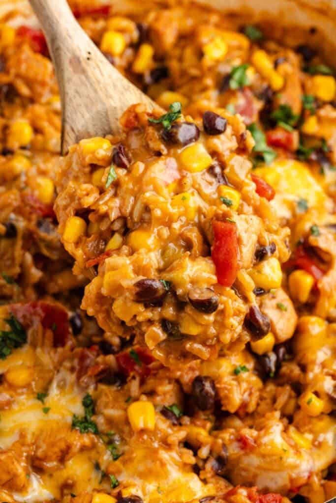 Wooden spoon scooping one-pot Mexican chicken and rice with black beans, corn, tomatoes, and herbs from a dish.