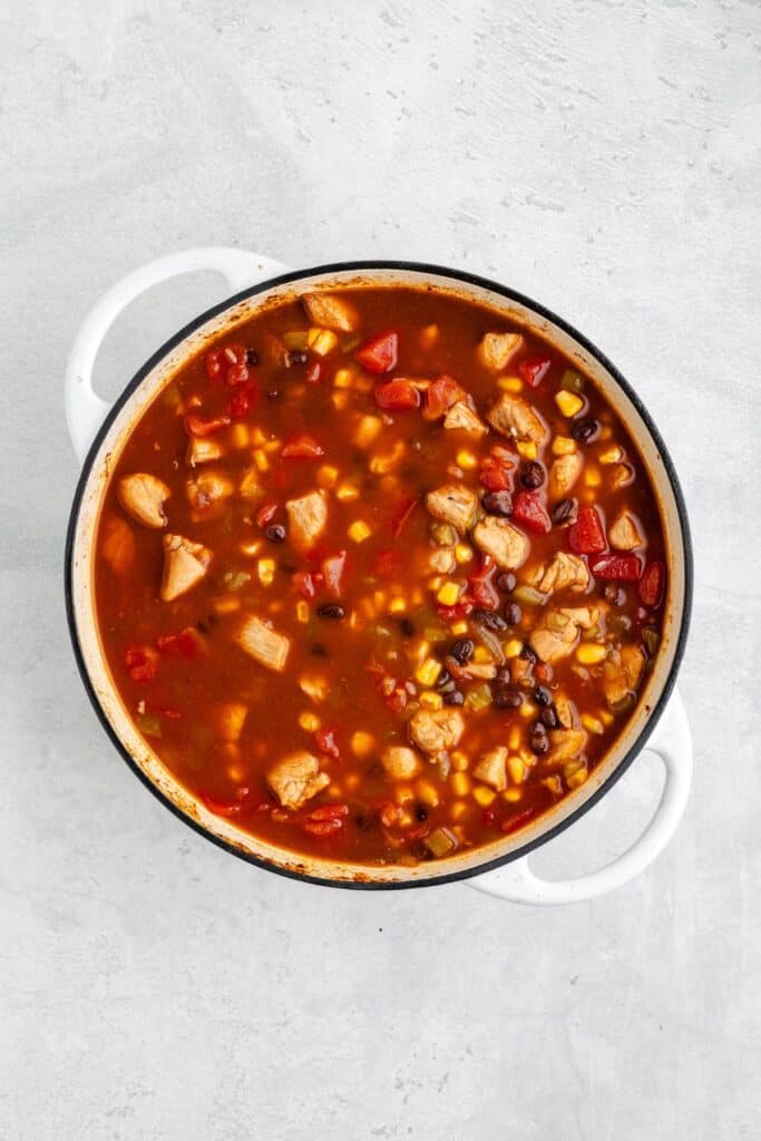 Chunky chicken soup with beans, corn, and veggies in a white pot, viewed from above on a light surface.