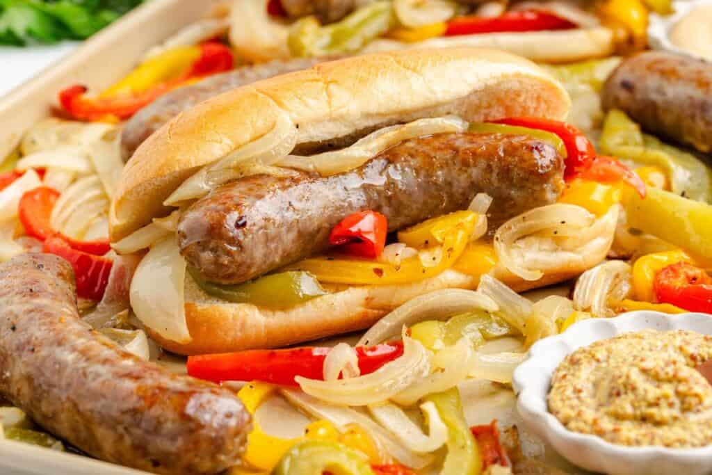 Sheet pan brats and peppers in a bun with sautéed onions, surrounded by more brats, vegetables, and mustard.