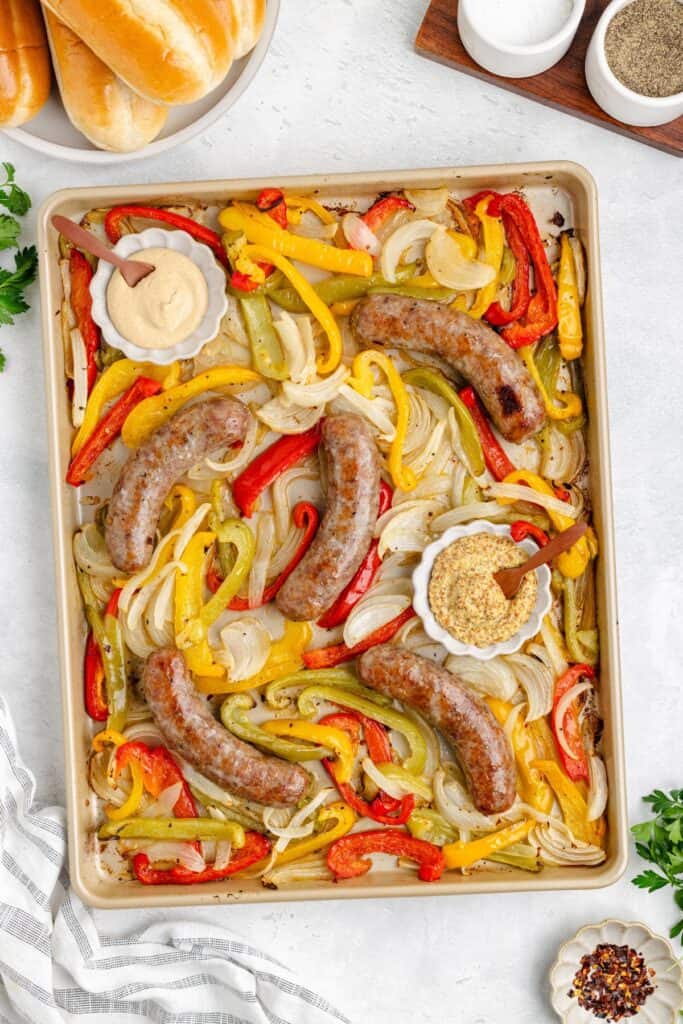 Sheet pan with cooked sausages, colorful bell peppers, onions, mustard and sauce bowls, plus buns and seasonings nearby.