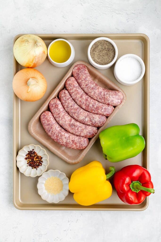 Uncooked sausages with bell peppers and onions, seasoned and placed on a baking sheet, ready for roasting.