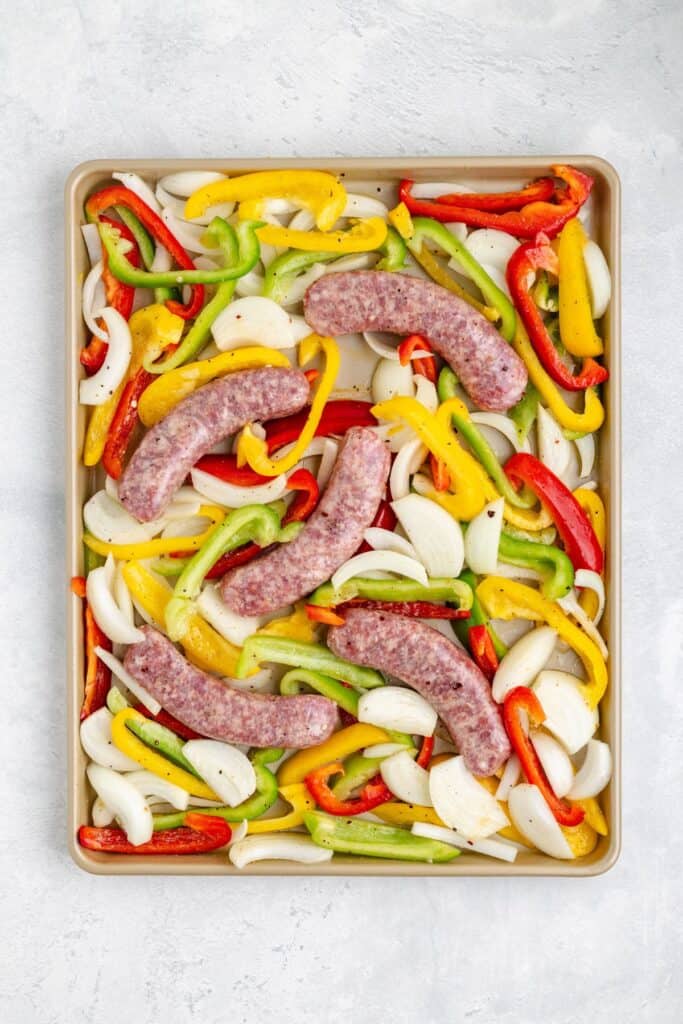Raw sausages, sliced bell peppers, and onions arranged on a baking sheet, ready to be roasted.