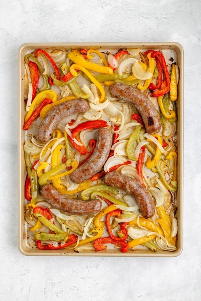 Sheet pan of cooked sausages with sliced red and yellow bell peppers and onions on a light background.